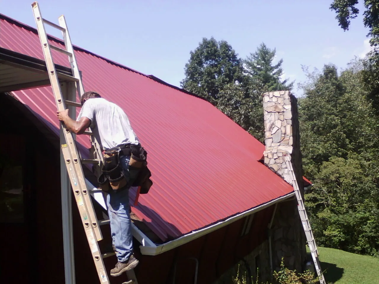 Roofer on a ladder beside a red metal roof and stone chimney — Soffit & Fascia Repair by Roofing Brothers in Placerville